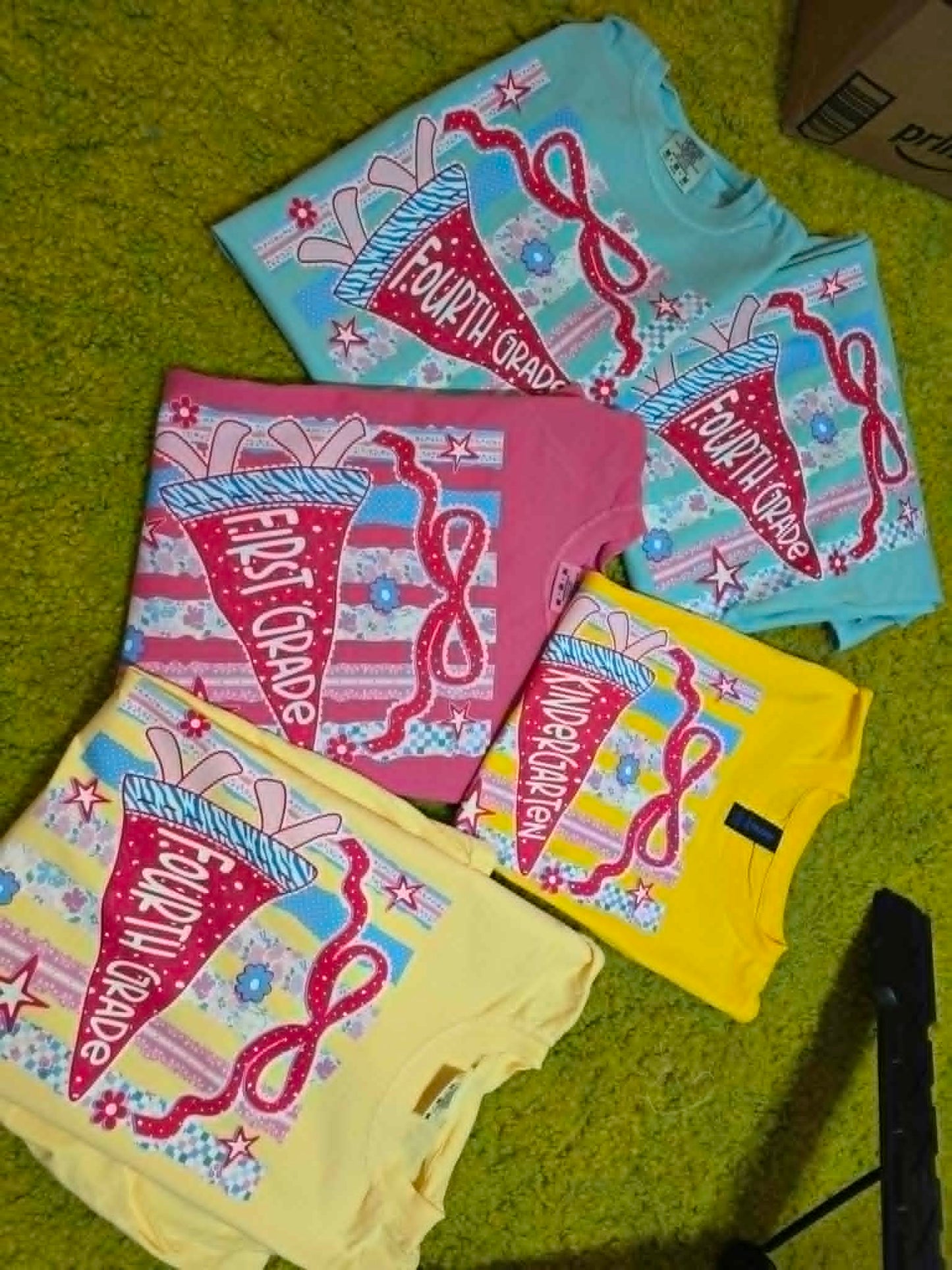 Colorful grade-level shirts with school pennants, pencils, bows, and classroom-themed patterns
