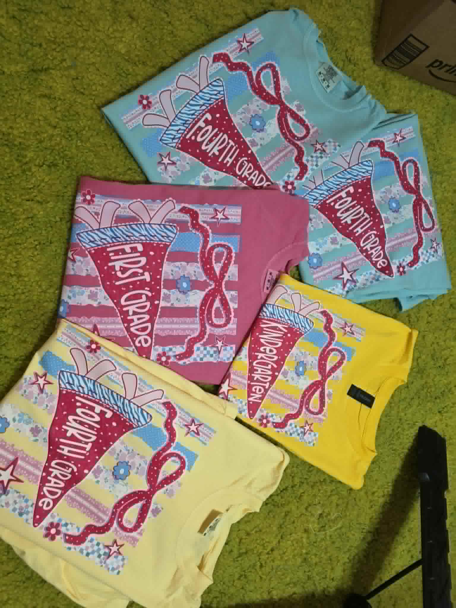 Colorful grade-level shirts with school pennants, pencils, bows, and classroom-themed patterns